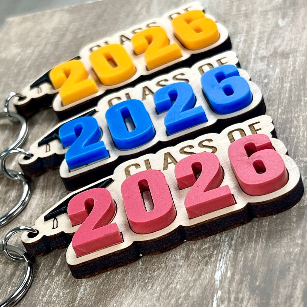 Multiple Class of 2026 maple graduation keychains with acrylic grad caps displaying mango, blue and watermelon numeral colour variants