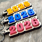 Multiple Class of 2026 maple graduation keychains with acrylic grad caps displaying mango, blue and watermelon numeral colour variants