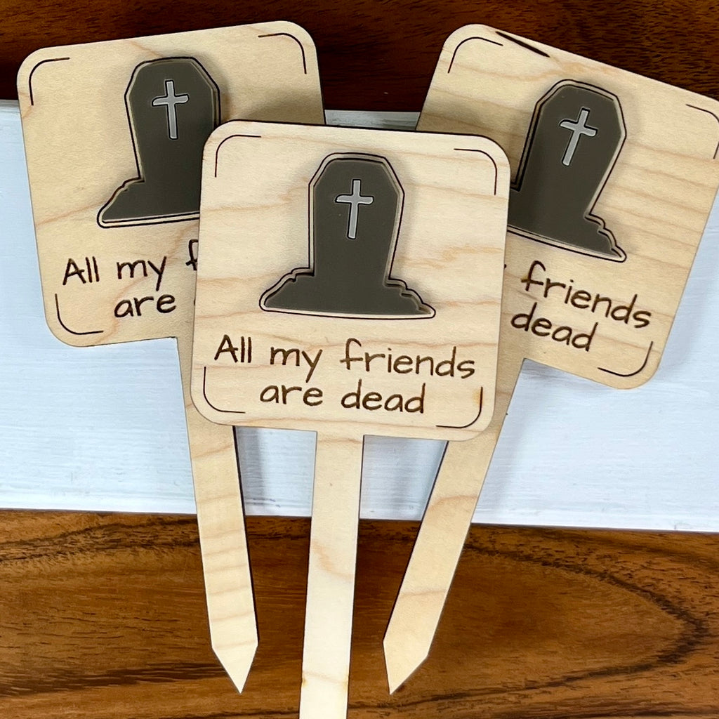Three wooden plant markers, each displaying a layered acrylic headstone icon and the phrase 'All my friends are dead' on a light wood background.