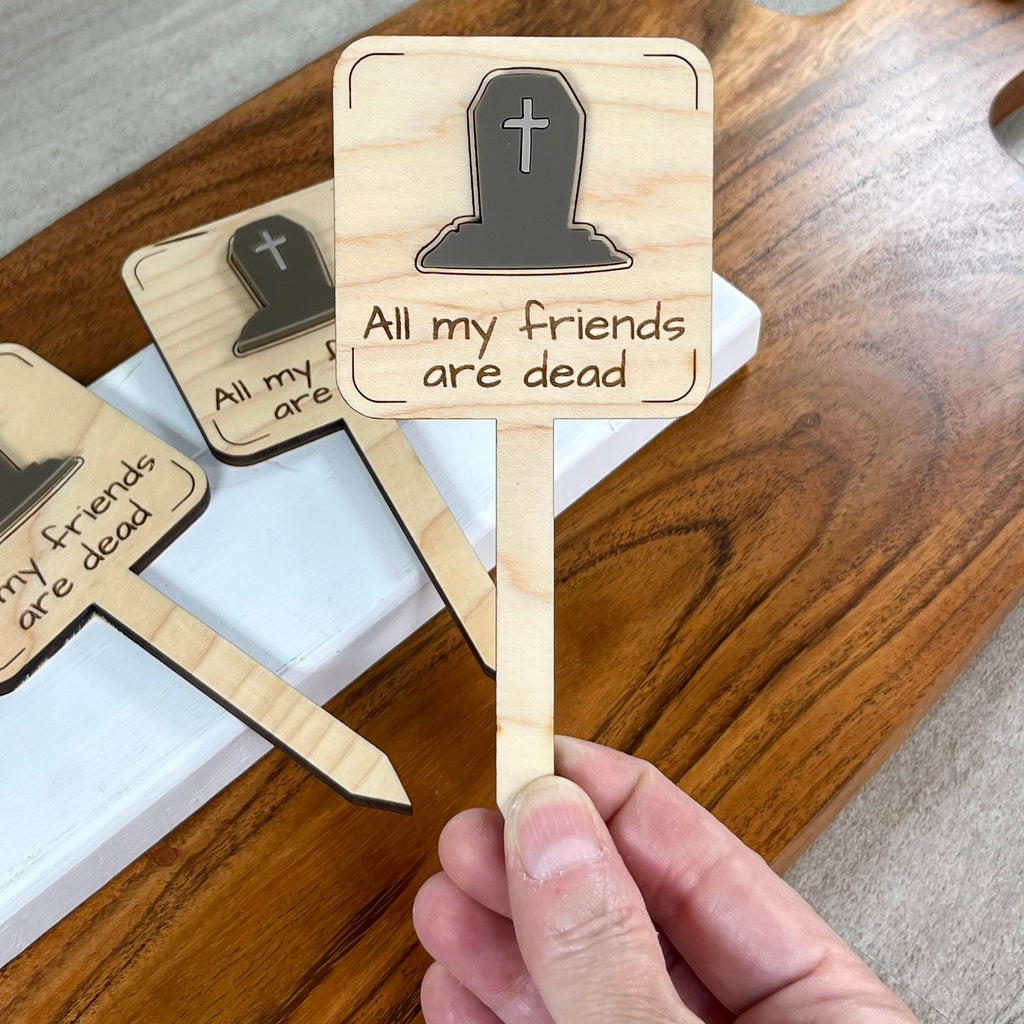 A hand holds a wooden plant stake displaying a layered acrylic headstone and the engraved text 'All my friends are dead' with more matching stakes on a wooden tray in the background.