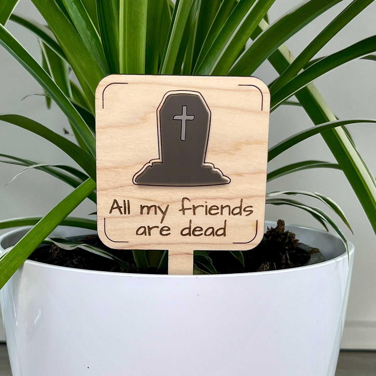 A square wooden garden stake with an acrylic headstone icon and the laser-engraved text 'All my friends are dead', displayed in a white planter pot with green plant leaves in the background.