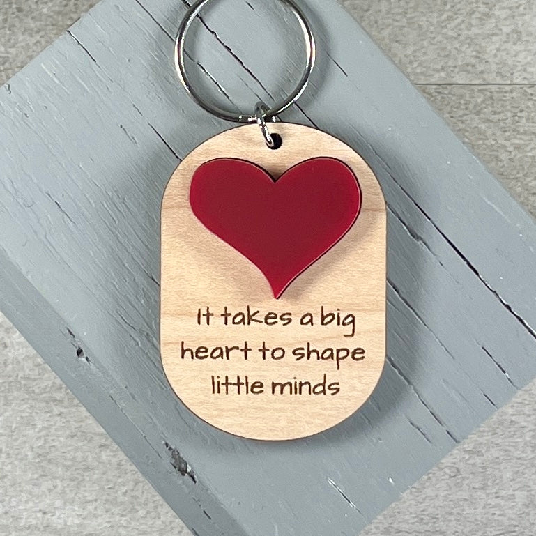 A wooden dog-tag style keychain with a red heart and the laser-engraved phrase 'It takes a big heart to shape little minds,' shown on a distressed gray wooden block.