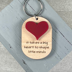 A wooden dog-tag style keychain with a red heart and the laser-engraved phrase 'It takes a big heart to shape little minds,' shown on a distressed gray wooden block.