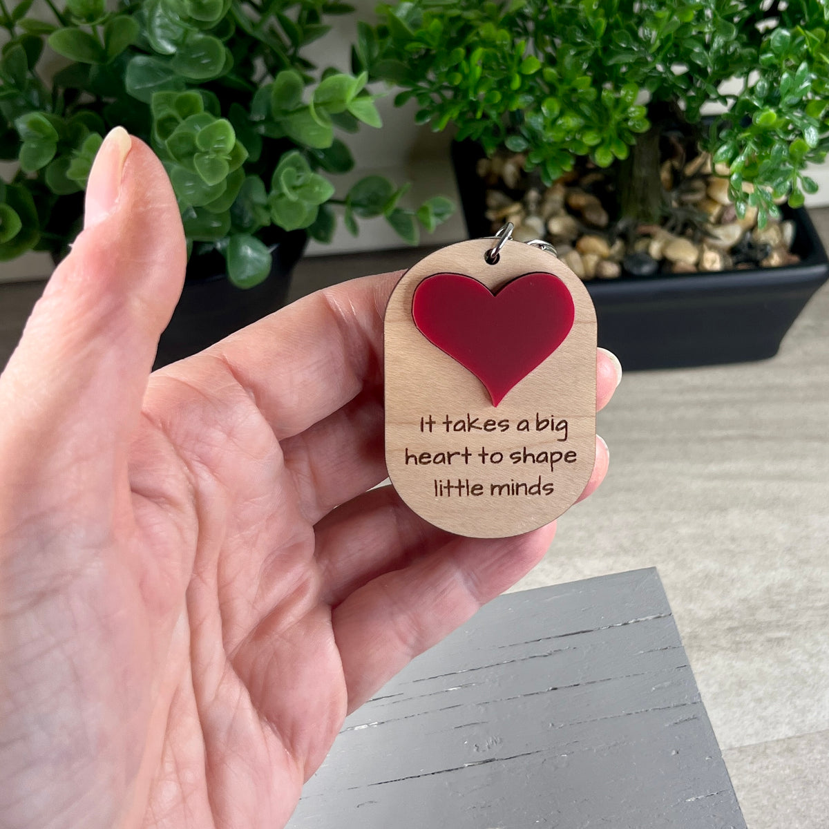 A hand holds a small oval wooden keychain with a red heart and the laser-engraved phrase 'It takes a big heart to shape little minds' against a blurred indoor background with potted green plants.