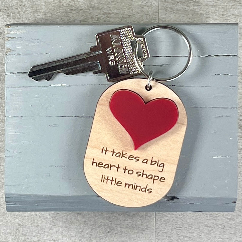 A wooden dog-tag style keychain with a red heart and laser-engraved quote, attached to a metal key on a distressed light blue wooden block.