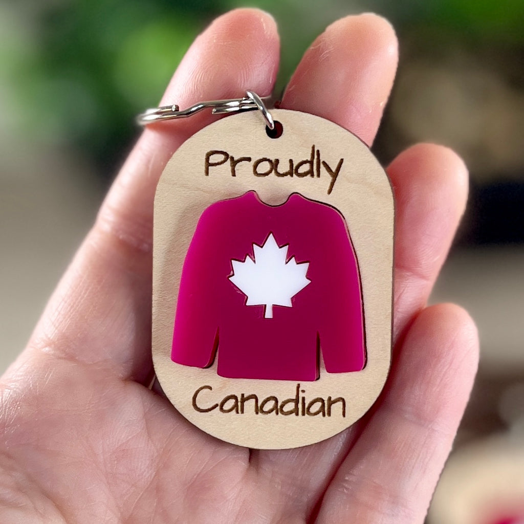 Hand holding Proudly Canadian maple keychain showing red acrylic Canadian hockey jersey detail