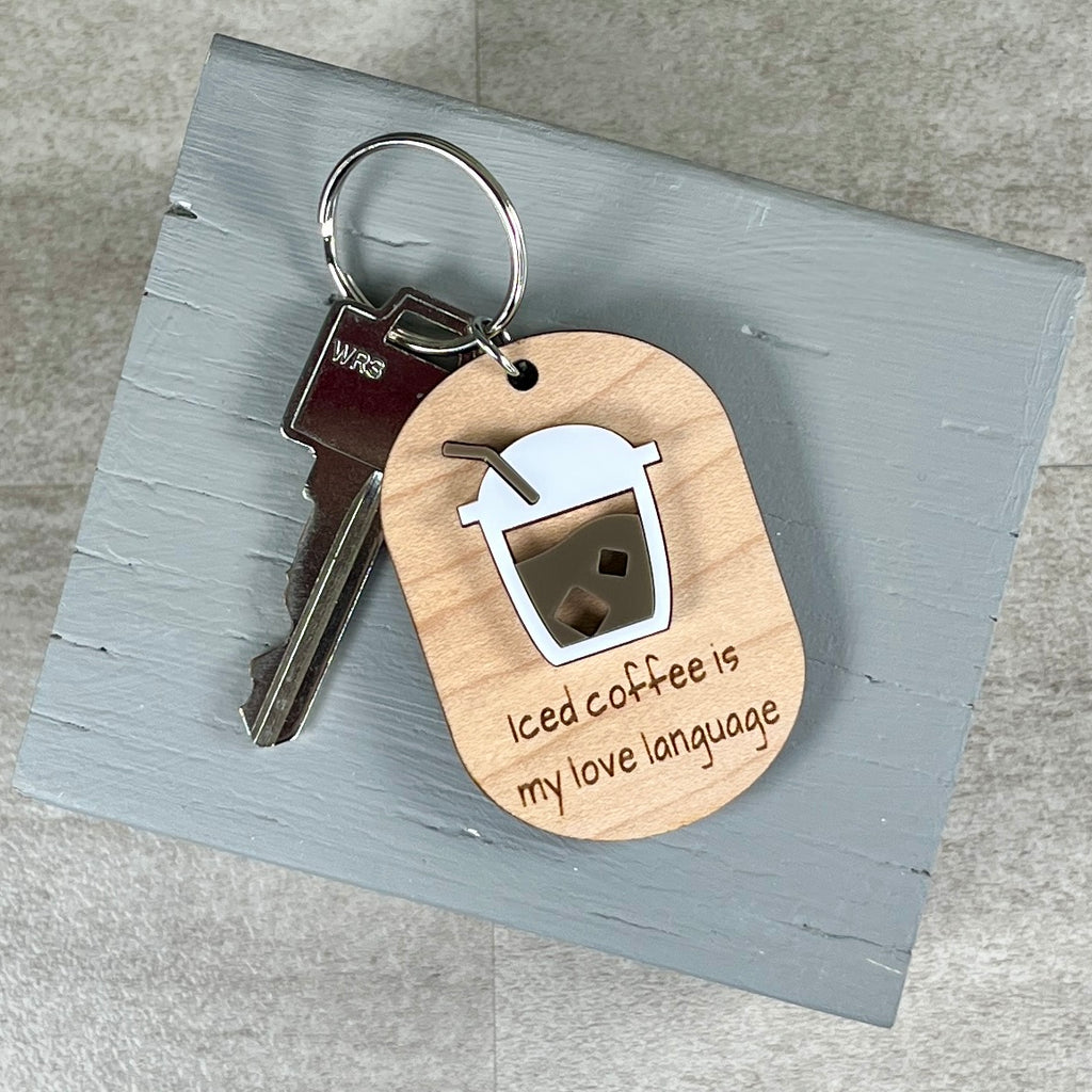 A wooden keychain with a metal key attached, featuring a layered acrylic coffee cup and the laser-engraved phrase 'Iced coffee is my love language' on a light gray wooden block