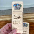 Hand holding Current Status Ignoring Responsibilities birch bookmark showing overflowing acrylic laundry basket