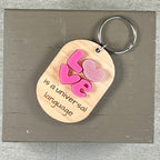 Oval wooden keychain with pink acrylic LOVE letters and engraved 'is a universal language,' with a metal split ring.