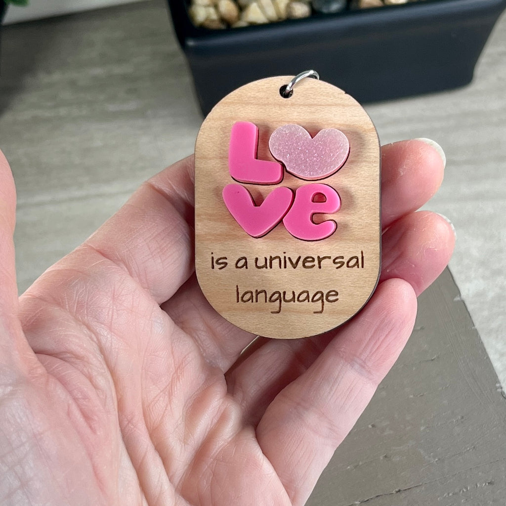 A hand holding an oval wooden keychain featuring layered pink acrylic LOVE letters above the engraved phrase 'is a universal language.'