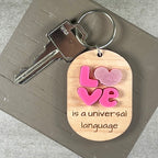 Oval wooden keychain with pink acrylic LOVE letters and engraved 'is a universal language,' with a metal split ring.