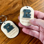 Hand holding I'd Rather Be Baking maple keychain showing grey acrylic stand mixer detail