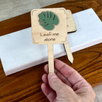 Hand holding two Leaf Me Alone laser-cut maple wood plant stakes with sage green monstera leaf