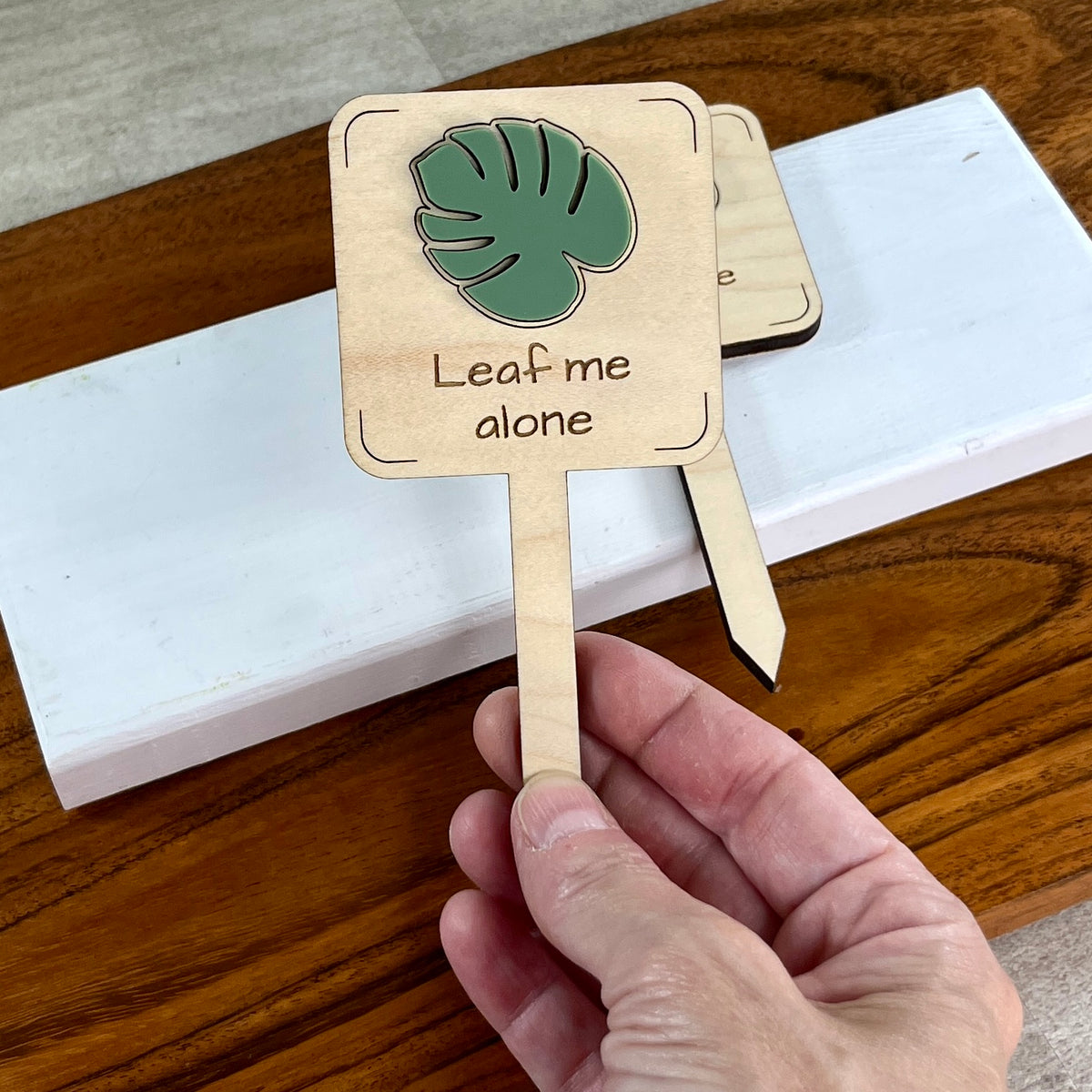 Hand holding two Leaf Me Alone laser-cut maple wood plant stakes with sage green monstera leaf