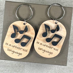 Two oval wooden keychains each featuring black acrylic musical notes above the laser-engraved phrase 'is a universal language' - the notes represent the word Music.