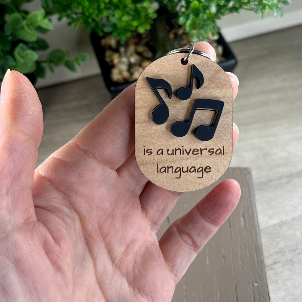A handheld oval wooden keychain with black acrylic musical notes above the laser-engraved phrase 'is a universal language' - the notes represent the word Music.