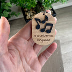 A handheld oval wooden keychain with black acrylic musical notes above the laser-engraved phrase 'is a universal language' - the notes represent the word Music.