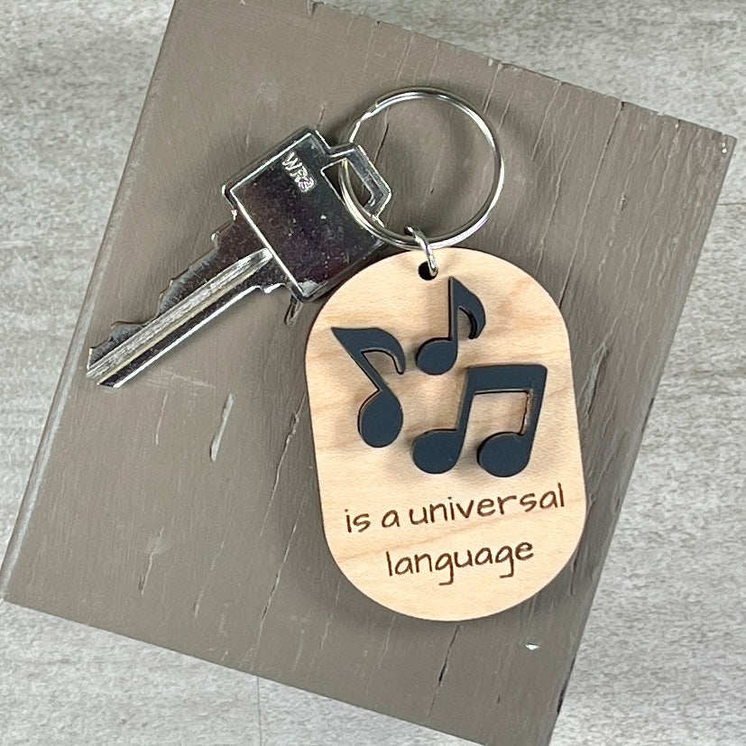 An oval wooden keychain with black acrylic musical notes above the phrase 'is a universal language,' with a key on a gray wood block over a light gray surface.