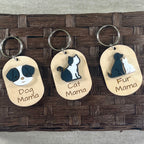 Three oval pet-tag style keychains with metal key rings are displayed on a brown woven mat. Each tag features a different pet illustration and the engraved text 'Dog Mama', 'Cat Mama', and 'Fur Mama'.