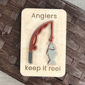 A small maple magnet with a layered acrylic fishing rod with a fish on the line, with the laser-engraved message “Anglers keep it reel,” placed on a brown woven surface.