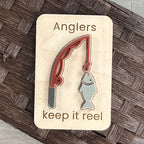 A small maple magnet with a layered acrylic fishing rod with a fish on the line, with the laser-engraved message “Anglers keep it reel,” placed on a brown woven surface.
