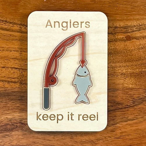 A small maple magnet with a layered acrylic fishing rod with a fish on the line, with the laser-engraved message “Anglers keep it reel,” placed on a brown wooden surface.