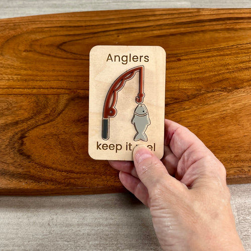 A hand holding a small wooden magnet featuring a layered acrylic fishing rod and fish design with the laser-engraved words 'Anglers keep it reel', set against a polished wooden board background.