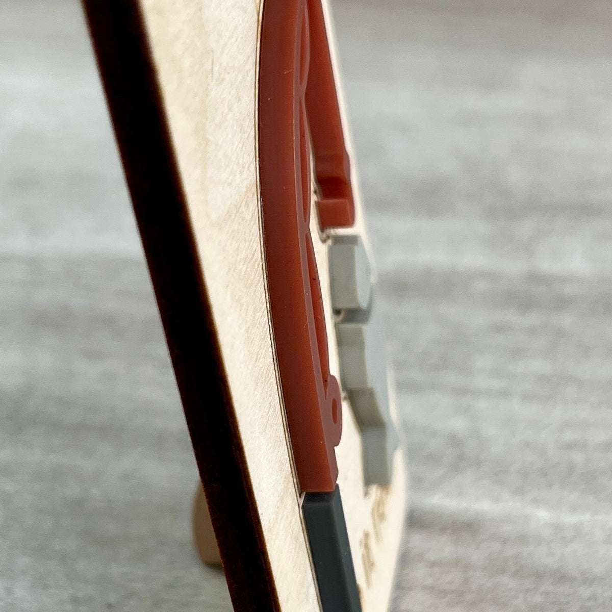 Side view of a wooden magnet with a light natural wood base, highlighting the 3D layered reddish-brown acrylic fishing rod, standing on a small round wooden base against a gray textured background.