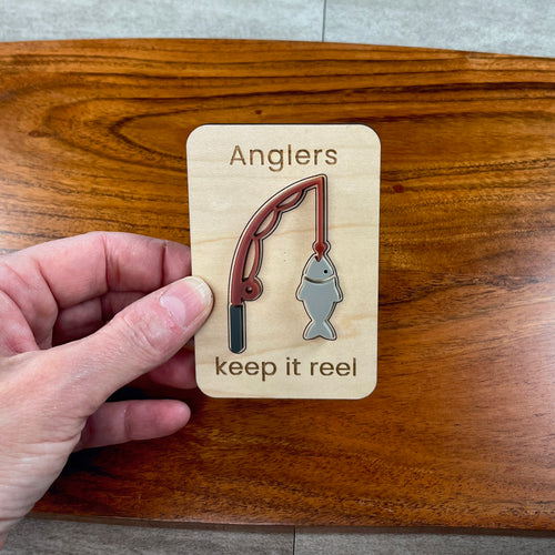 A hand holding a small wooden magnet featuring a layered acrylic fishing rod and fish design with the laser-engraved words 'Anglers keep it reel', set against a polished wooden board background.