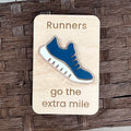 A rectangular wooden magnet laser-engraved with the words 'Runners go the extra mile' and featuring a layered blue and white acrylic running shoe in the center, displayed on a brown woven mat background.
