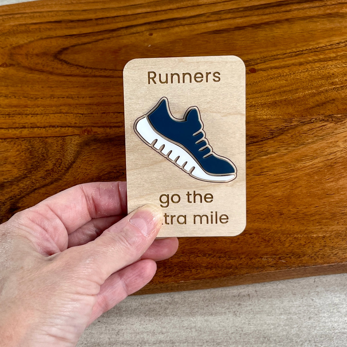 A hand holds a small wooden magnet laser-engraved with the words 'Runners go the extra mile,' featuring a layered navy blue and white acrylic running shoe in the center. The plaque is positioned over a polished wooden surface.