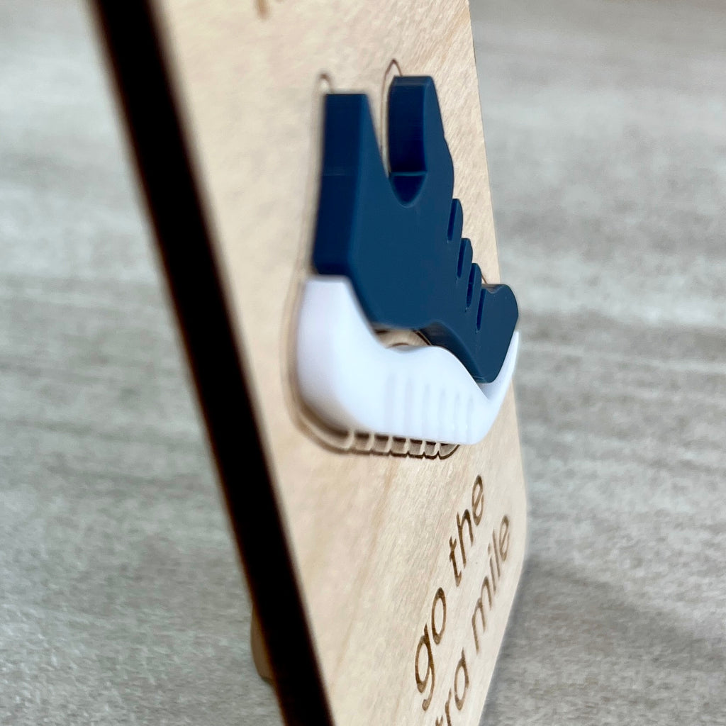 A side view close-up of a wooden magnet featuring a layered acrylic navy blue and white running shoe, with engraved text on the wood, set against a soft gray background designed to highlight the 3D effect of the acrylic layer.