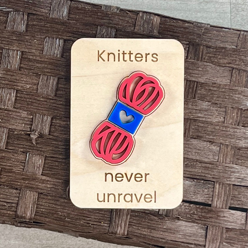 A small maple magnet layered with a pink and blue skein of yarn and laser-engraved with the message “Knitters never unravel,” placed on a brown woven surface.