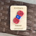 A small maple magnet layered with a pink and blue skein of yarn and laser-engraved with the message “Knitters never unravel,” placed on a brown woven surface.