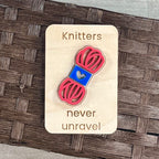 A small maple magnet layered with a pink and blue skein of yarn and laser-engraved with the message “Knitters never unravel,” placed on a brown woven surface.