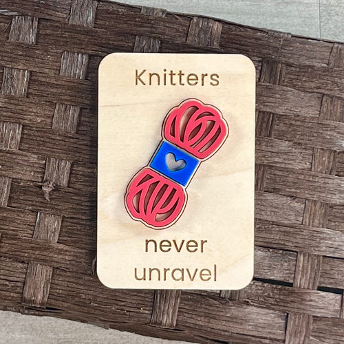 A small maple magnet layered with a pink and blue skein of yarn and laser-engraved with the message “Knitters never unravel,” placed on a brown woven surface.