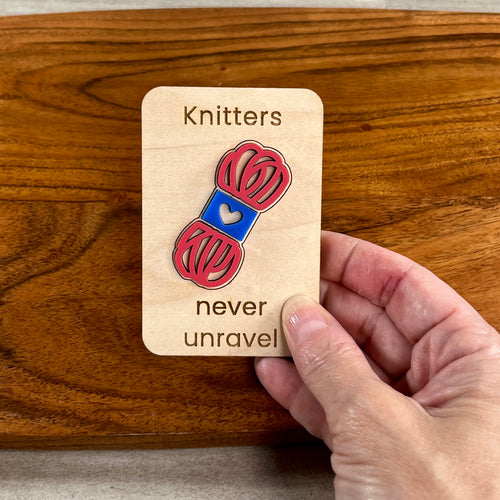 A hand holds a wooden magnet laser-engraved with the words 'Knitters never unravel', featuring a pink and blue acrylic skein of yarn held against a warm brown wooden background.