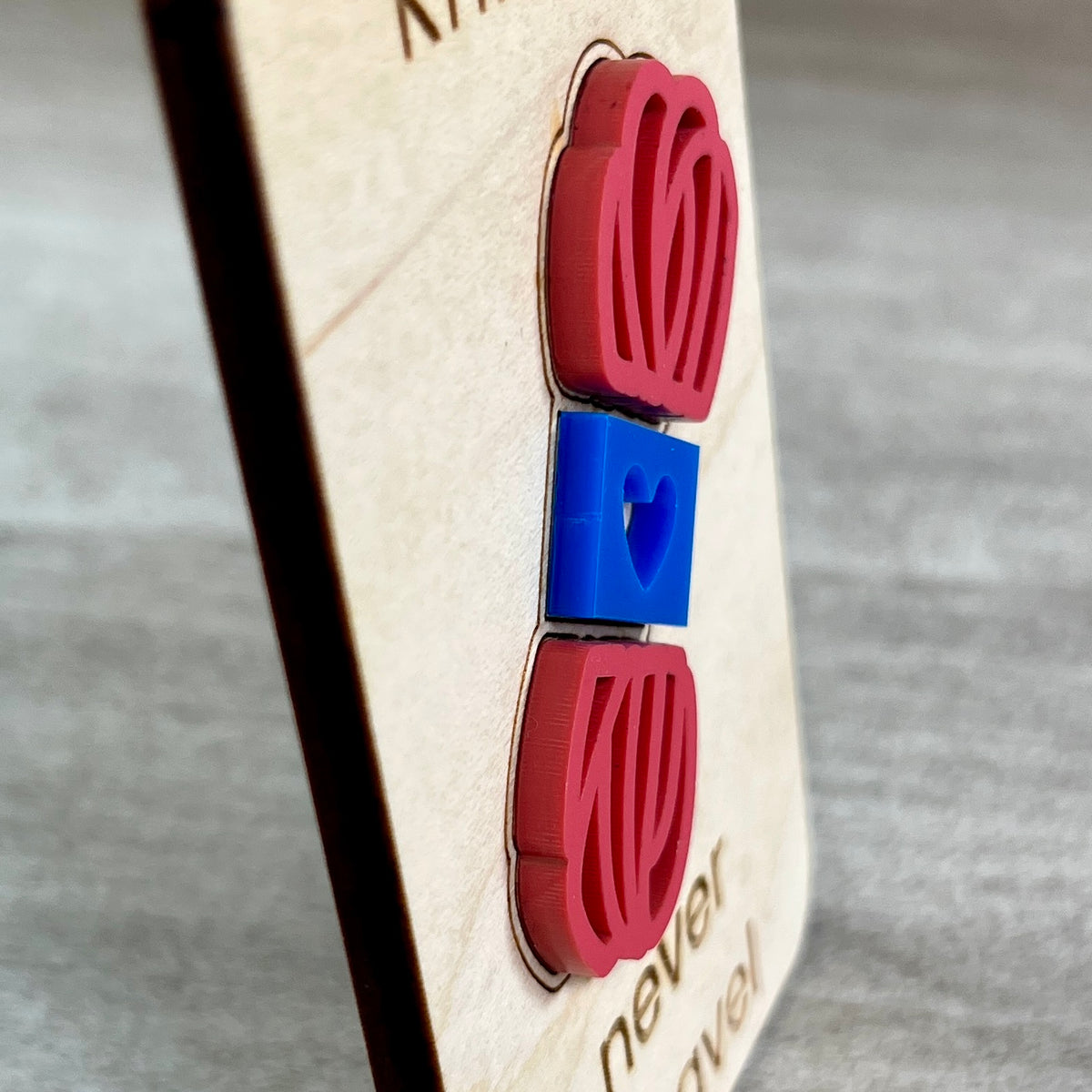 A side view close-up of a wooden magnet layered with a pink and blue acrylic skein of yarn, photographed against a gray textured background and designed to highlight the 3D effect of the yarn element.