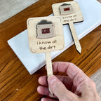 Hand holding two I Know All the Dirt laser-cut maple wood plant stakes with cream acrylic soil bag