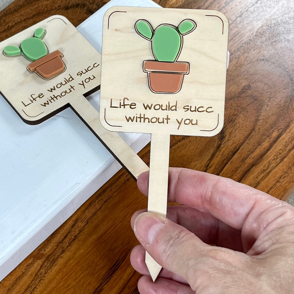 Hand holding two Life Would Succ Without You laser-cut maple wood plant stakes with green succulent