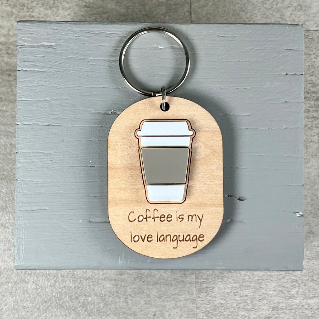 A wooden keychain with a metal split ring, featuring an engraved oval tag that reads 'Coffee is my love language' with an acrylic coffee cup design, displayed on a gray wooden block.