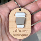 A hand holding a light wood oval keychain with an acrylic coffee cup illustration and the laser-engraved phrase 'Coffee is my love language'.