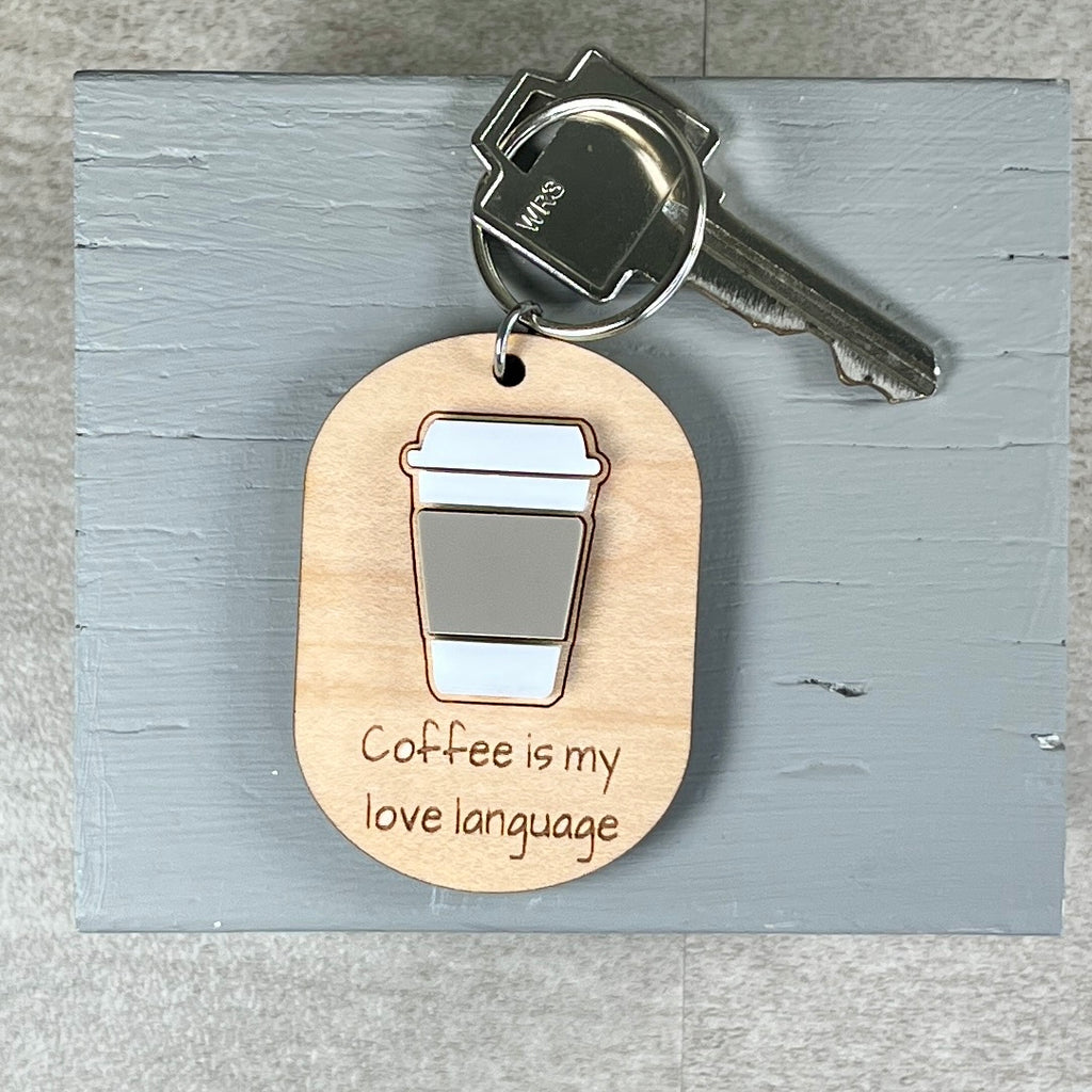 A wooden coffee-themed keychain with a metal key attached, featuring an acrylic coffee cup graphic and the engraved phrase 'Coffee is my love language' on a light gray wooden board.