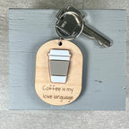 A wooden coffee-themed keychain with a metal key attached, featuring an acrylic coffee cup graphic and the engraved phrase 'Coffee is my love language' on a light gray wooden board.