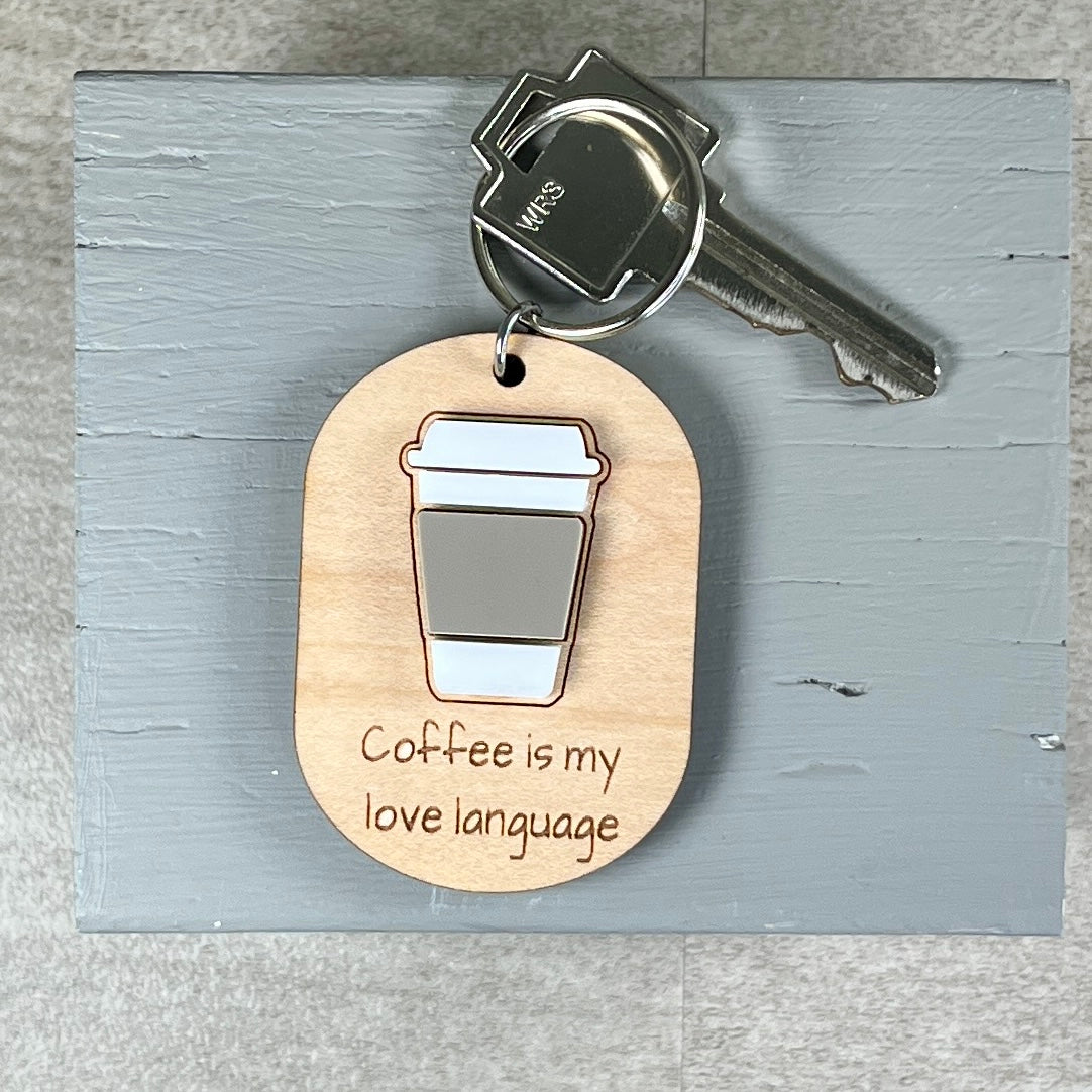A wooden coffee-themed keychain with a metal key attached, featuring an acrylic coffee cup graphic and the engraved phrase 'Coffee is my love language' on a light gray wooden board.