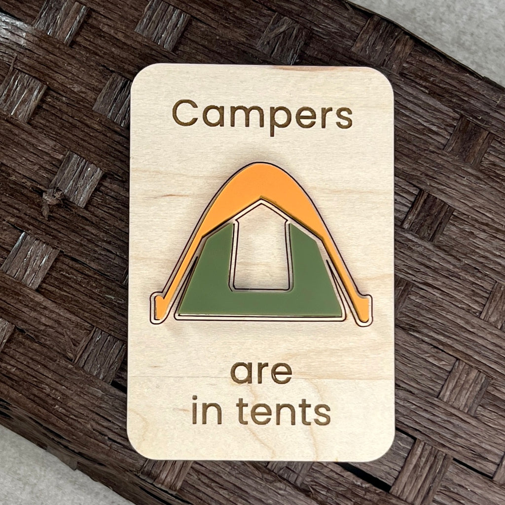 A rectangular wooden magnet with rounded corners featuring the laser-engraved text 'Campers are in tents' and a stylized layered acrylic tent in orange and green, displayed on a dark woven surface.