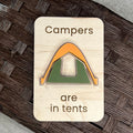 A rectangular wooden magnet with rounded corners featuring the laser-engraved text 'Campers are in tents' and a stylized layered acrylic tent in orange and green, displayed on a dark woven surface.