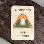 A rectangular wooden magnet with rounded corners featuring the laser-engraved text 'Campers are in tents' and a stylized layered acrylic tent in orange and green, displayed on a dark woven surface.