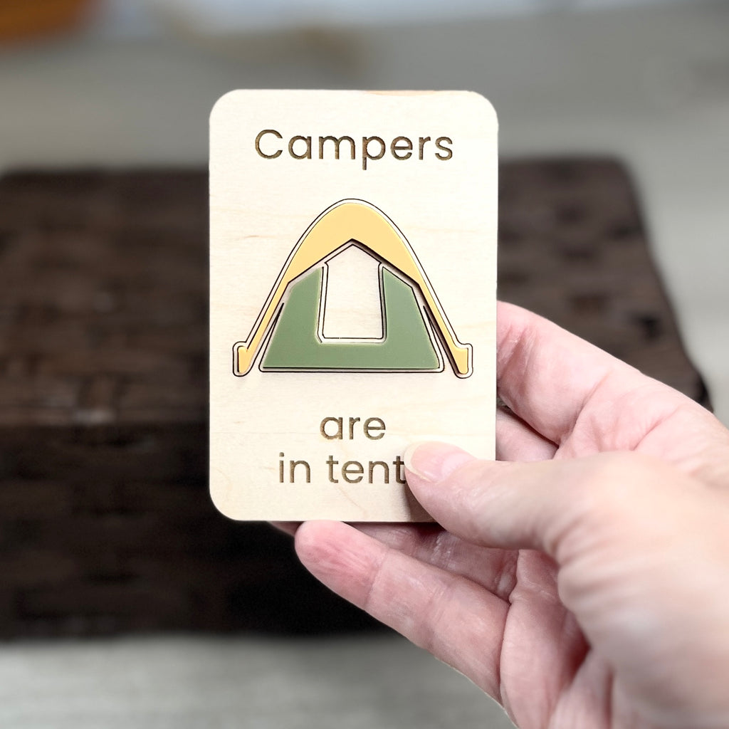 A hand holds a small wooden magnet with the laser-engraved message 'Campers are in tents' and a simple acrylic tent design. The background is softly blurred, showing a dark woven basket and light floor.