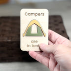 A hand holds a small wooden magnet with the laser-engraved message 'Campers are in tents' and a simple acrylic tent design. The background is softly blurred, showing a dark woven basket and light floor.